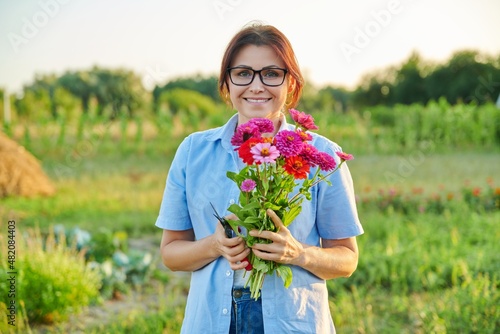 Smiling middle aged female holding bouquet of fresh zinnia flowers in garden