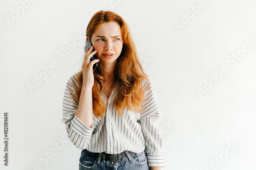 Confused redhead young woman talking with a mobile phone with her friend and frowning, isolated on white