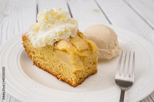 Piece of apple cake with biscuit base and whipped cream topping, served with vanilla ice cream close up on a plate on wooden background