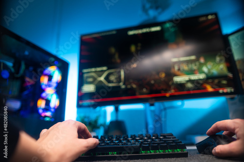 A gamer sits at the keyboard of a personal computer and plays a video game on a large monitor with friends. Neon lighting. Gaming, virtual reality, cybersport, modern technologies.