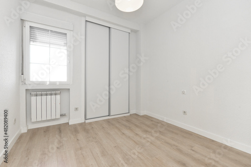 Empty room with light wooden floor, sliding white wooden wardrobe doors and aluminum window above aluminum radiator and ceiling lamp