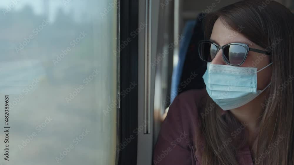 Woman in medical mask looking through the train window while traveling. Traveling during Covid. Woman in medical mask looking through the train window while traveling. Traveling during Covid.