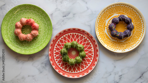 Three colorful plates with a geometric design, with three different Mochi Donuts.  Strawberry, Ube, and Matcha glaze.