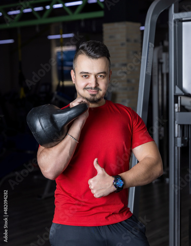 Athletic man in a red shirt holding kettlebell in his hand for strength workout at the gym