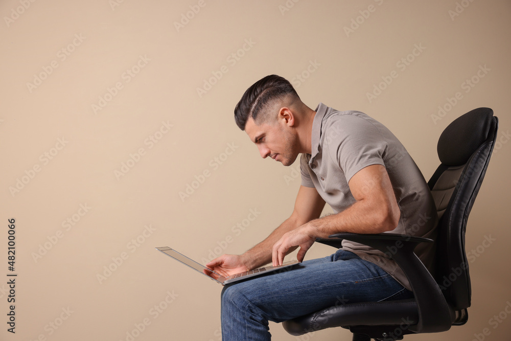 Man with poor posture using laptop while sitting on chair against beige background, space for text Man with poor posture using laptop while sitting on chair against beige background, space for text