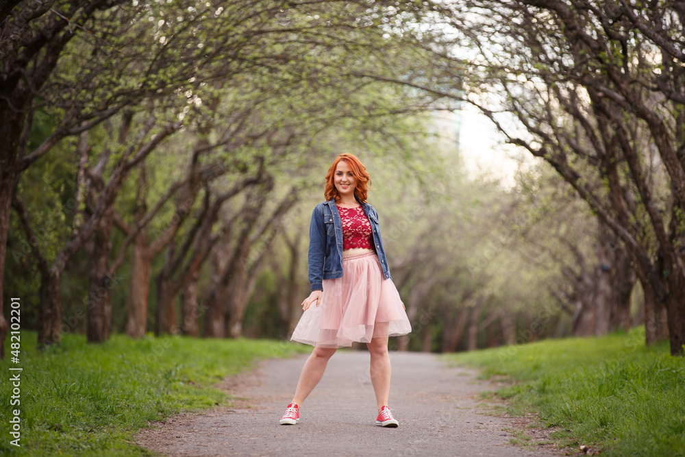 Active and happy woman in spring park. Young redhaired girl on background of green trees lane. Redhaired girl in peach skirt and jeans jacket and sneakers walks in nature. Young early spring Active and happy woman in spring park. Young redhaired girl on background of green trees lane. Redhaired girl in peach skirt and jeans jacket and sneakers walks in nature. Young early spring