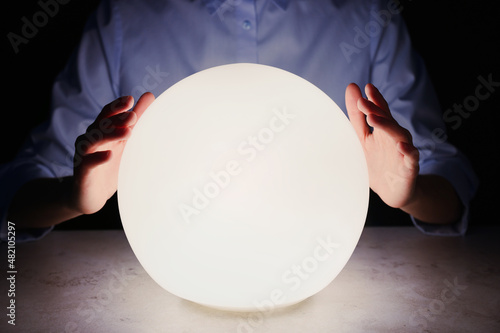 Businesswoman using glowing crystal ball to predict future at table in darkness, closeup. Fortune telling