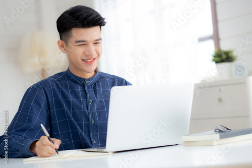 Young asian business man writing on notebook for planning working and using laptop computer on desk at home, notes about finance, male study and learning, business and communication concept.