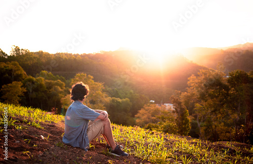 Young man sitting on top of a hill and watching the sunset. Person thinking and having reflections