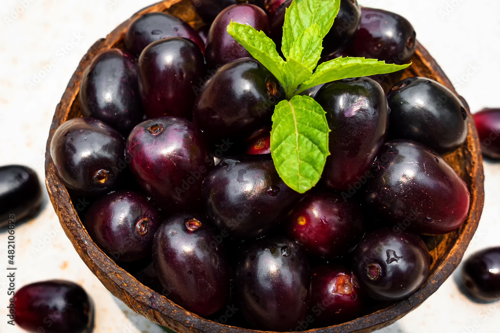 Dark pink-red ripe Syzygium cumini fruits. Dark black java plum in a wood bowl at isolated white background. Green mint leaf on top of some large java plums. Dark pink-red ripe Syzygium cumini fruits. Dark black java plum in a wood bowl at isolated white background. Green mint leaf on top of some large java plums.