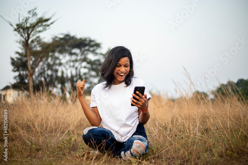 Full length Portrait of a excited beautiful black skinned African woman typing on the smart phone in a park with dry grass