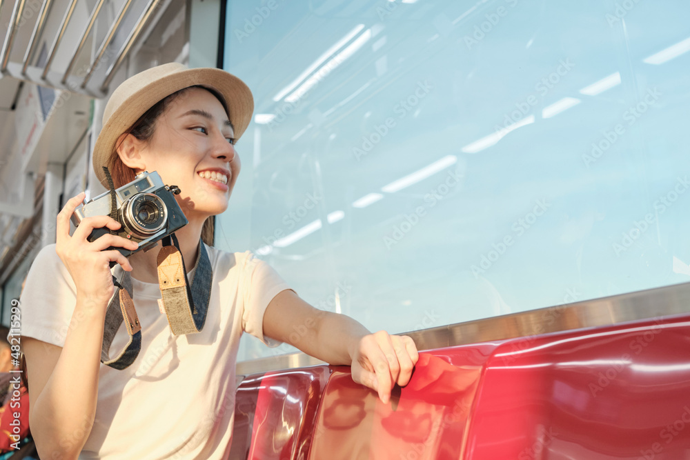 Naklejka premium Beautiful Asian female tourist sits in a red seat, traveling by train, taking snapshot photo, transporting in suburb view, enjoy passenger lifestyle by railway, happy journey vacation.