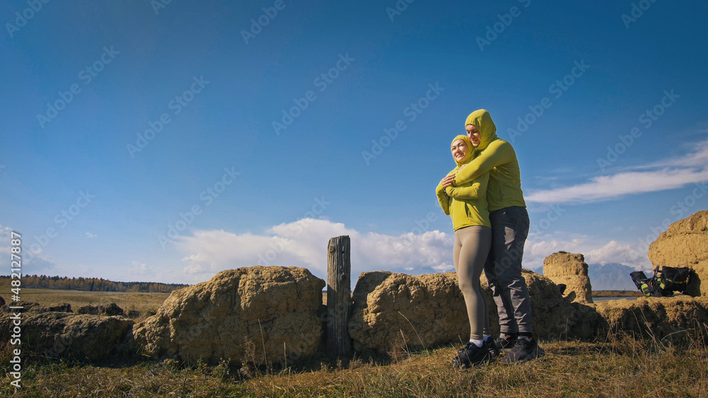 Man and woman in yellow green sportswear. Lovely couple of travelers hug and kiss near old stone enjoying highland landscape.