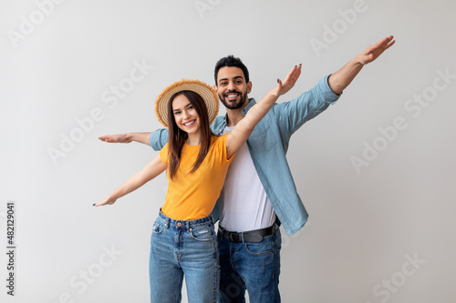 Young couple in love pretending airplane flight, spreading hands like wings, having fun while posing over light studio wall