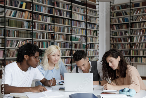 Multiethnic group of four gen Z university students working together at laptop, writing notes, watching learning webinar, online lecture, reading tutorials on internet in college library