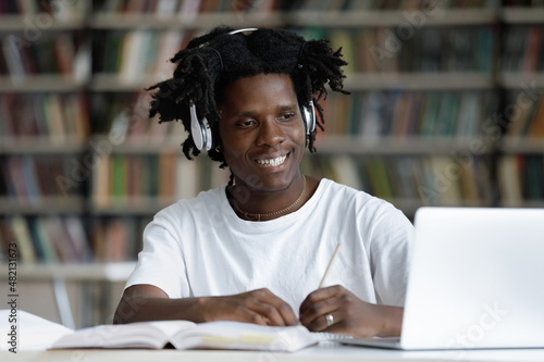 Happy gen Z African student guy in headphones watching learning webinar, online lesson on laptop, writing notes, consulting teacher on video call on internet, working on study project in library