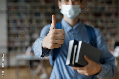 Young man making thumb up hand close up. College student guy wearing medical facial mask visiting public place, auditorium, university library, recommending protection from infecton during epidemic photo
