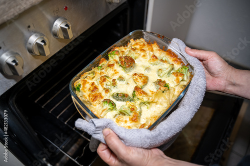 Woman holds baking pan with pasta, broccoli and cheese gratin in front of the oven