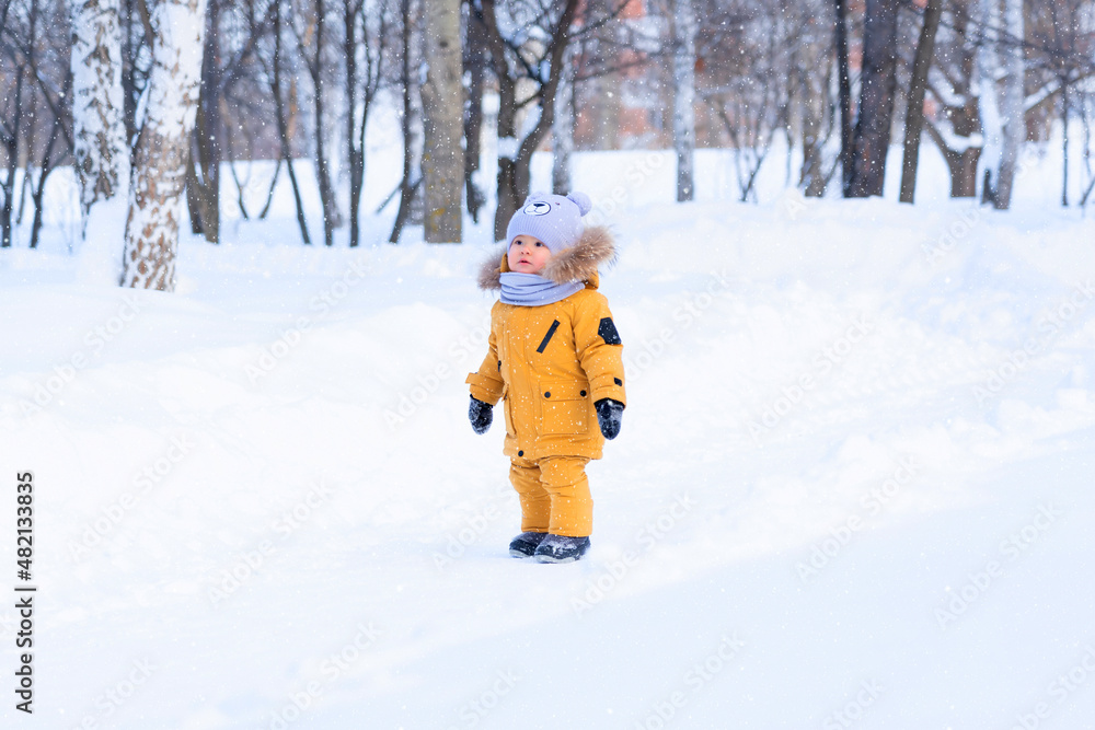 Portrait of a toddler 15-20 months old in yellow warm clothes in a winter park looks at the falling snowflakes with surprise