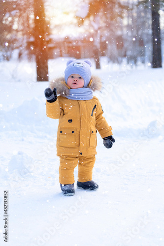 The kid greets by raising his hand and looking forward. Portrait of a toddler 15-20 months old in yellow warm clothes during a snowfall.