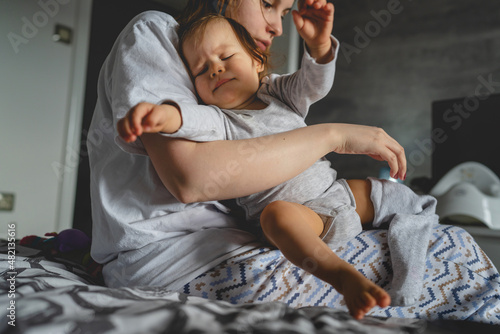 One small caucasian baby girl and her mother morning routine getting dressed woman having hard time trouble while putting on clothes on her nine months old daughter real people family copy space