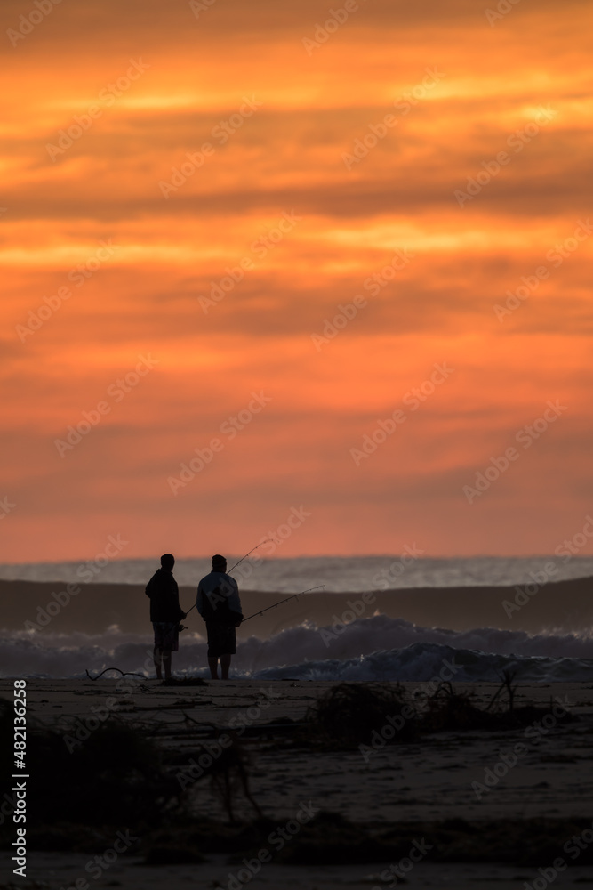 Fishermen standing on a surf beach at sunrise with an orange sky.