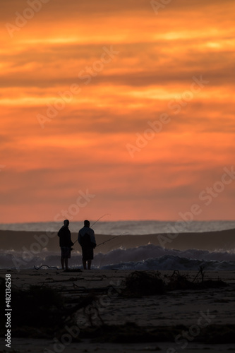 Fishermen standing on a surf beach at sunrise with an orange sky.