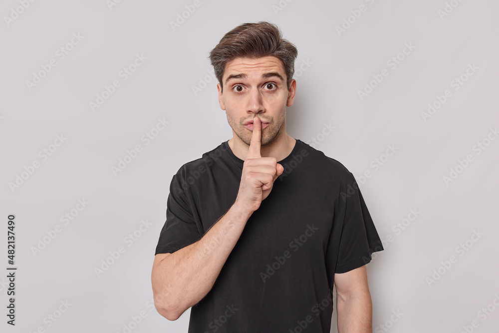 Surprised European place finger on lips asks to be quiet demans silence wears casual basic t shirt wants to hide secret isolated over white background. People body language and secrecy concept Surprised European place finger on lips asks to be quiet demans silence wears casual basic t shirt wants to hide secret isolated over white background. People body language and secrecy concept