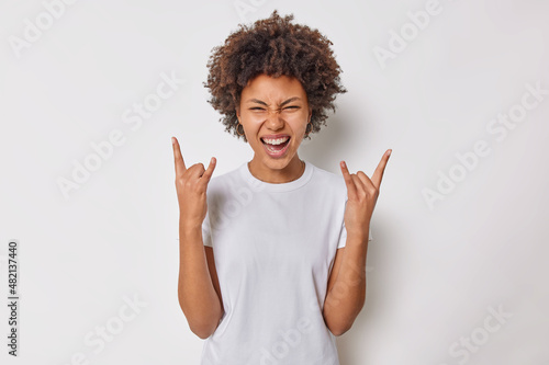 Carefree joyful woman with curly hair makess rock n roll gesture heavy metal sign enjoys favorite music dressed casually isolated over white background spends free time on party. Body language photo