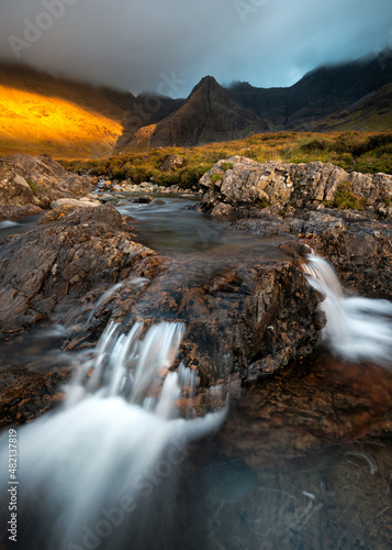 Fairy Pools Waterfall with view of famous Cuillin mountain range. Beautiful golden evening light with dark and dramatic clouds. Isle of Skye, Scotland, UK. © _Danoz