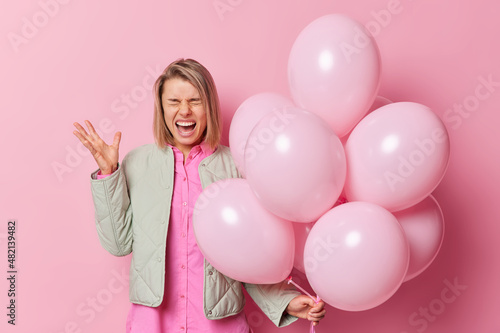 Emotional woman screams outraged shouts at someone holds bunch of inflated balloons keeps hand raised wears shirt and jacket expresses negative emotions angry to hear annoying music on party