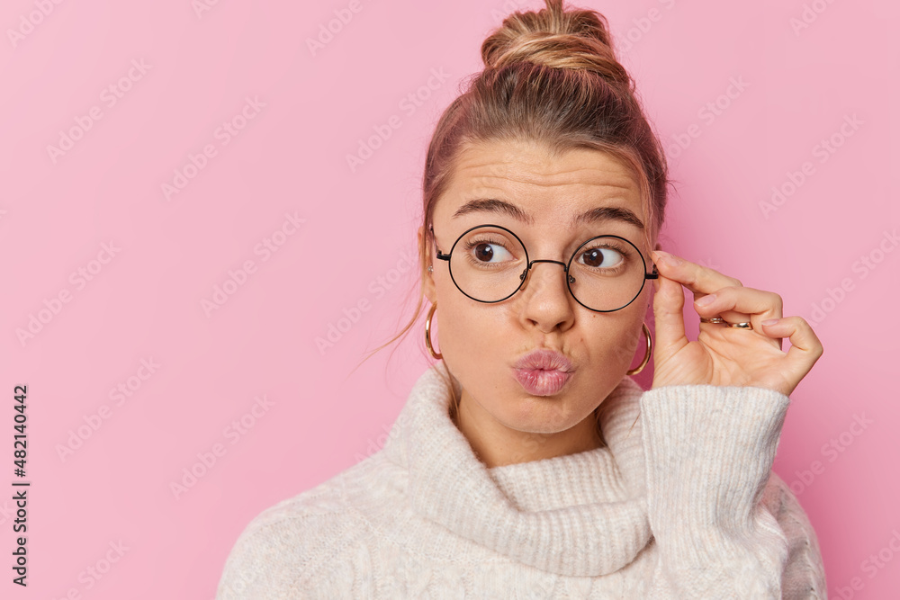 Portrait of good looking woman keeps lips rounded hand on rim of spectacles concentrated away with thoughtful expression dressed in warm sweater isolated over pink background considers something Portrait of good looking woman keeps lips rounded hand on rim of spectacles concentrated away with thoughtful expression dressed in warm sweater isolated over pink background considers something
