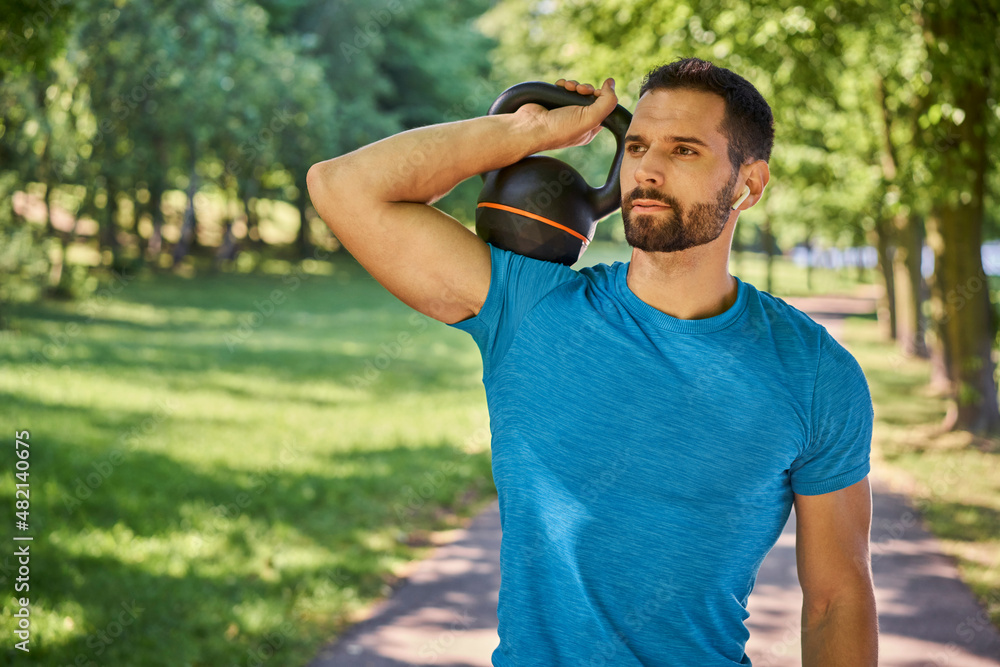 Obraz premium Portrait of an athletic man holding a kettlebell and looking away in a park