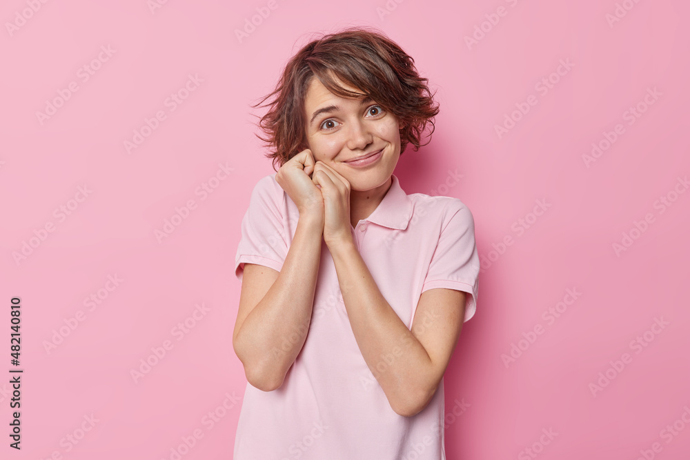 Portrait of charming young woman with short dark hair keeps hands near face wears casual t shirt poses against pink background. Joyful European female model hears pleasant news stands indoor Portrait of charming young woman with short dark hair keeps hands near face wears casual t shirt poses against pink background. Joyful European female model hears pleasant news stands indoor