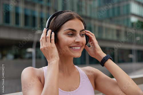 Positive dark haired woman puts on headphones smiles positively wears t shirt and smartwatch on wrist has glad expression poses against blurred background. People and healthy lifestyle concept