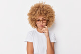 Photo of serious curly haired woman purses lips looks displeased being discontent with something keeps finger near lips wears spectacles and t shirt isolated over white background. Let me think