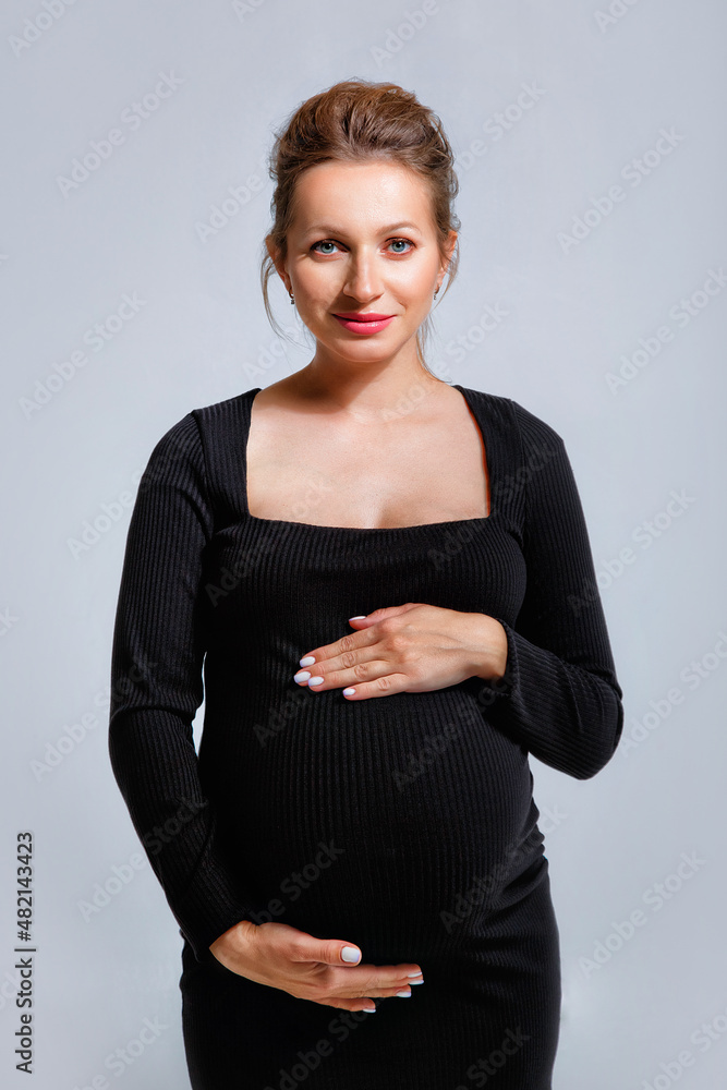 Portrait of a pregnant woman in a tight black dress on a gray background. The woman gently holds her stomach with her hands. Portrait of a pregnant woman in a tight black dress on a gray background. The woman gently holds her stomach with her hands.