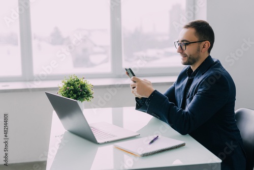 Slika na platnu Business man sitting at desk in his office, holding phone and smiling happily wh
