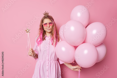 Displeased irritated birthday girl has image of princess wears crown sunglasses and dress holds magic wand and bunch of balloons being on party isolated over pink background. Celebration concept