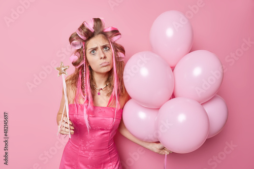 Displeased birthday girl prepares for celebration has leaked makeup after crying has some troubles looks disappointed applies hair curlers for making hairstyle holds bunch of balloons and magic wand