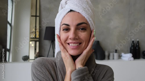 A young woman at home in the bathroom in a bathrobe with a towel on her head admires herself, looks at the camera as if in a mirror, touches her face with her fingers 