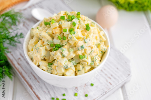 Egg salad with chopped green onions on top in a white bowl for cooking a sandwich on a white wooden background. Selective focus