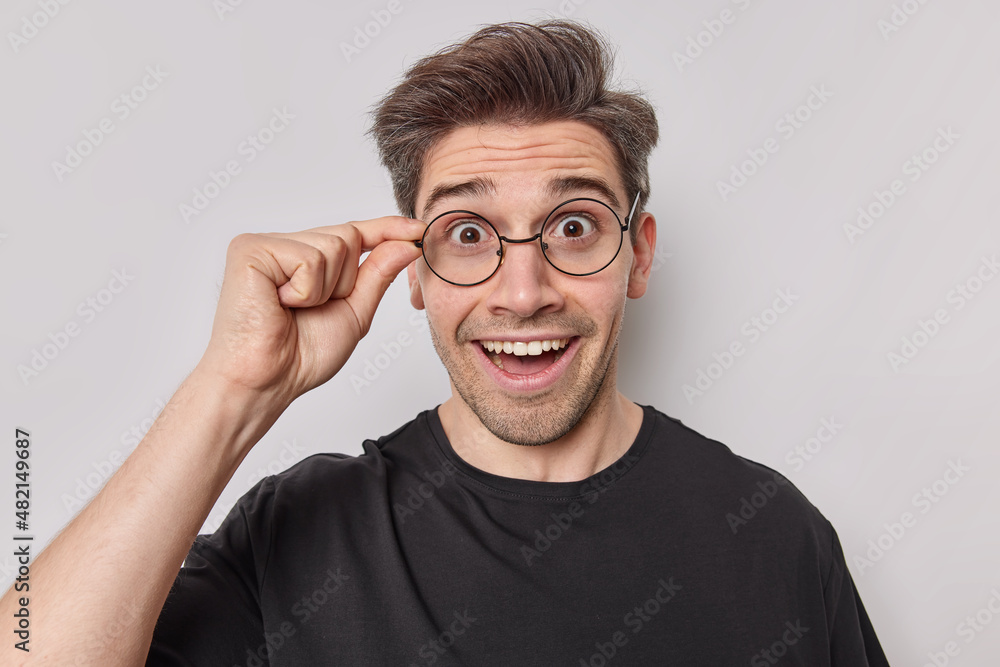 Surprised cheerful adult Caucasian man looks with joy at awesome promo keeps hand on rim of spectacles reacts on something isolated over white background looks amazed. Human reactions concept Surprised cheerful adult Caucasian man looks with joy at awesome promo keeps hand on rim of spectacles reacts on something isolated over white background looks amazed. Human reactions concept