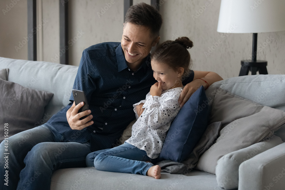 Joyful young caring father and adorable laughing small kid daughter looking at cellphone screen, making funny video, posing for selfie photo, playing entertaining games, modern tech addiction concept. Joyful young caring father and adorable laughing small kid daughter looking at cellphone screen, making funny video, posing for selfie photo, playing entertaining games, modern tech addiction concept.