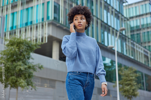 Serious beautiful young woman with curly hair wears casual jumper and jeans calls someone via smartphone has conversation poses against modern city building. People lifestyle technology concept