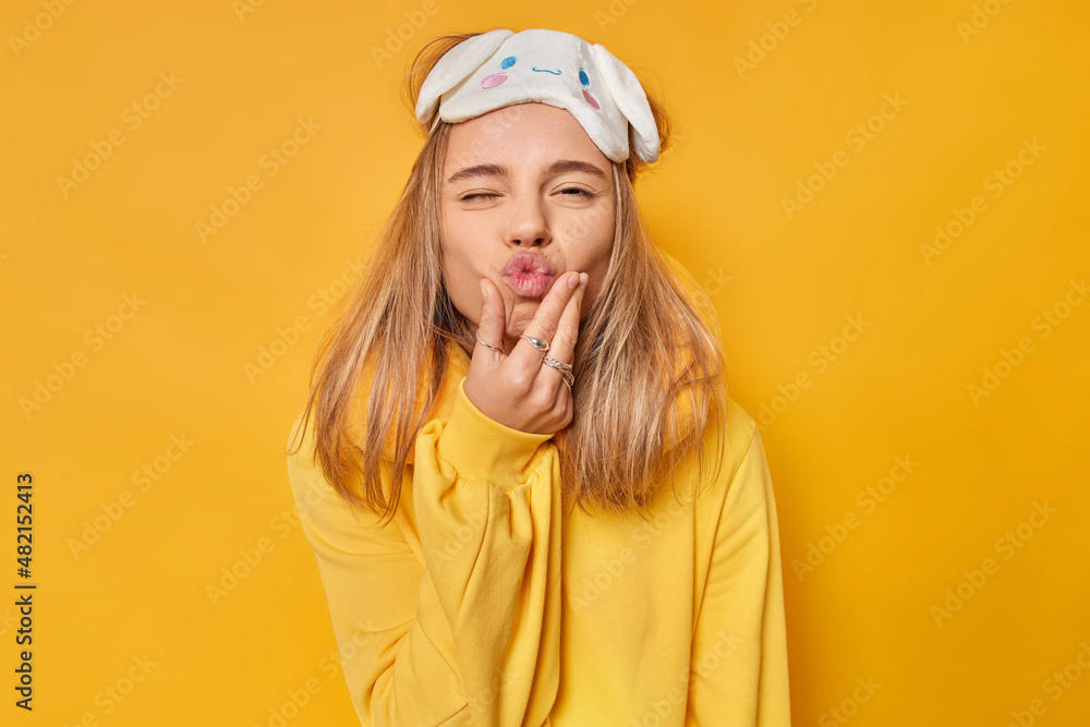 Funny sleepy woman keeps lips rounded wears blindfold neckpillow and casual jumper wants to kiss you poses against vivd yellow background. People rest sleeping and facial expressions concept Funny sleepy woman keeps lips rounded wears blindfold neckpillow and casual jumper wants to kiss you poses against vivd yellow background. People rest sleeping and facial expressions concept