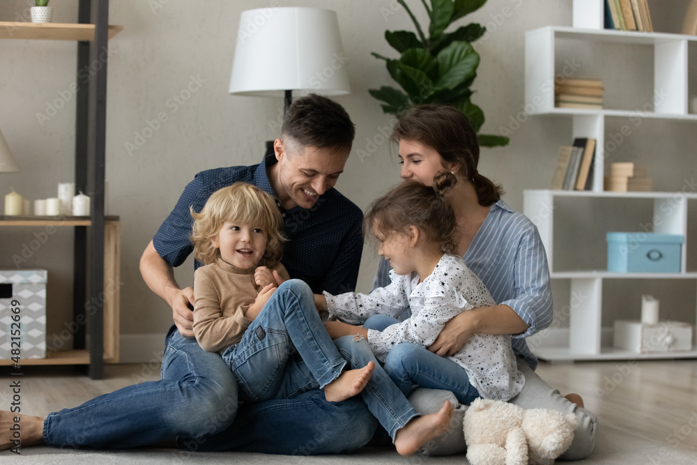 Smiling affectionate young couple parents sitting on floor carpet, holding on laps joyful sincere little preschool children siblings, having fun playing enjoying carefree weekend time together at home