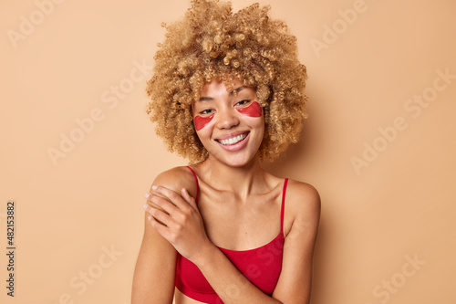 Portrait of cheerful curly haired young woman keeps hand on shoulder wears red cropped top applies hydrogel patches under eyes for skin treatment isolated over beige background. Beauty concept