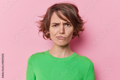 Portrait of young displeased woman frowns face has sulking expression purses lips has short hairstyle wears green jumper isolated dover pink studio background. Negative human emotions concept
