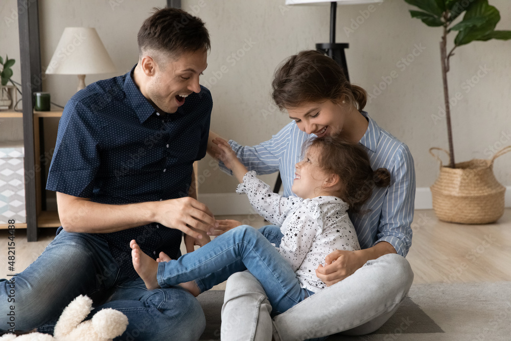 Laughing bonding young couple parents tickling happy adorable little kid daughter, having fun together on floor carpet, spending leisure weekend time together at home, family relations concept. Laughing bonding young couple parents tickling happy adorable little kid daughter, having fun together on floor carpet, spending leisure weekend time together at home, family relations concept.
